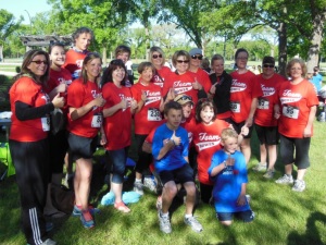 A team of individuals wearing red tshirts that say 'Team Bowes' give the thumbs up to the camera. Kneeling in front are two boys wearing blue shirts that say 'TeamHD'. The individuals are in a park with vivid green grass and large trees.